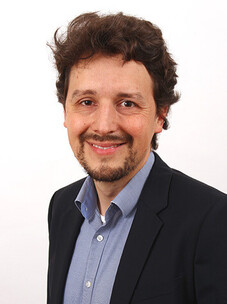 Lukas Janssen im Anzug, freundlich lächelnd vor hellem Hintergrund.